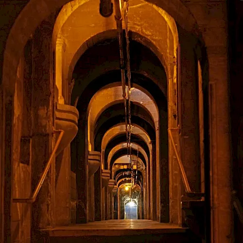A long, dim industrial tunnel with repeating arched openings, metal pipes, and warm glowing lights along the corridor.
