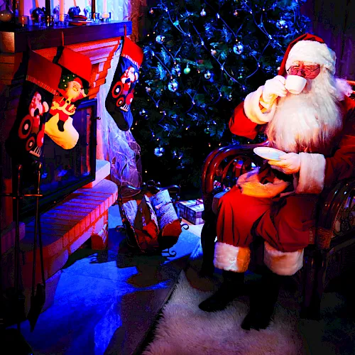 Santa in a red suit sits by a glowing fireplace, sipping a drink, with a festive tree and hanging ornaments behind him.