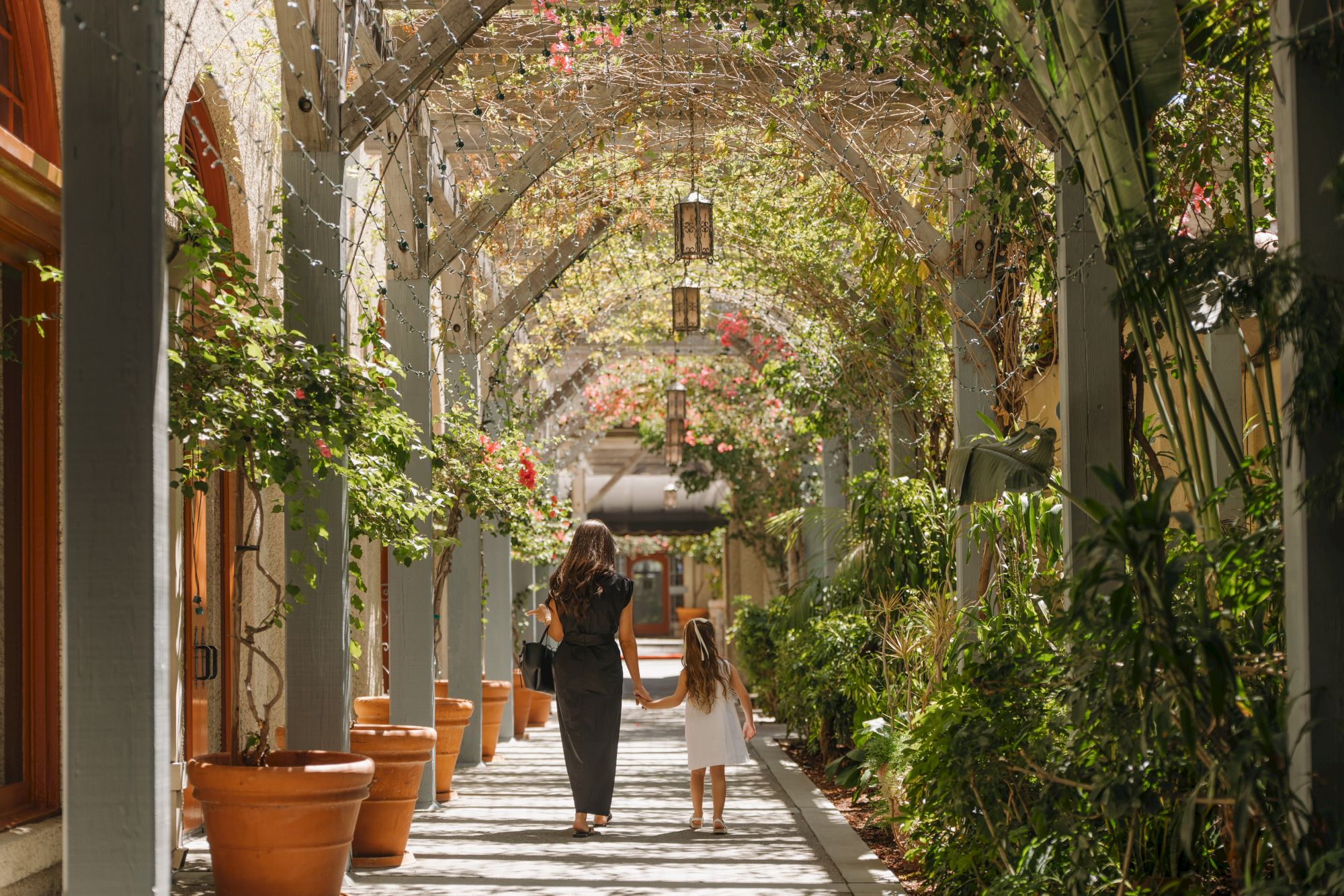 A person walks down a lush garden courtyard lined with potted plants, arches, and warm sunlit stone walls.