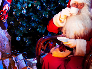 Santa in a red suit reads a book while holding his hands near his face, surrounded by gifts and blue lights in the background.