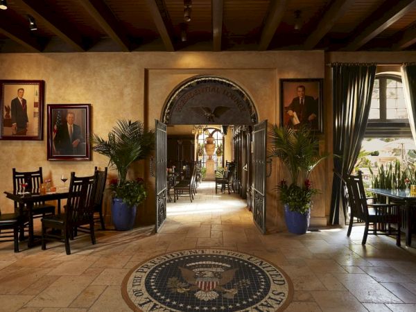 An elegant lounge corridor with framed portraits, potted palms, tables and chairs, and a central eagle emblem rug.