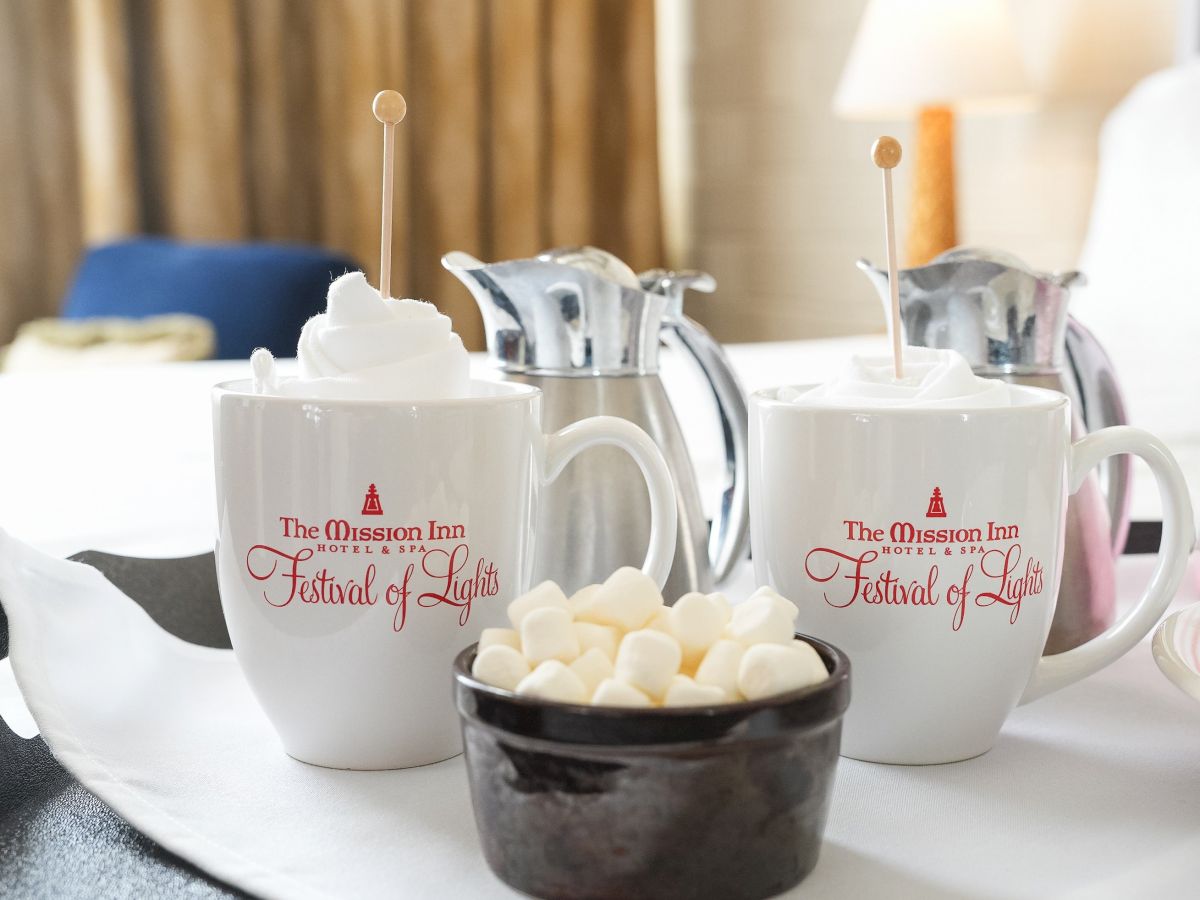 Two &ldquo;The Mission Inn&rdquo; mugs with stir sticks sit by a teapot on a table, with sweets for the &ldquo;Festival of Lights.&rdquo;