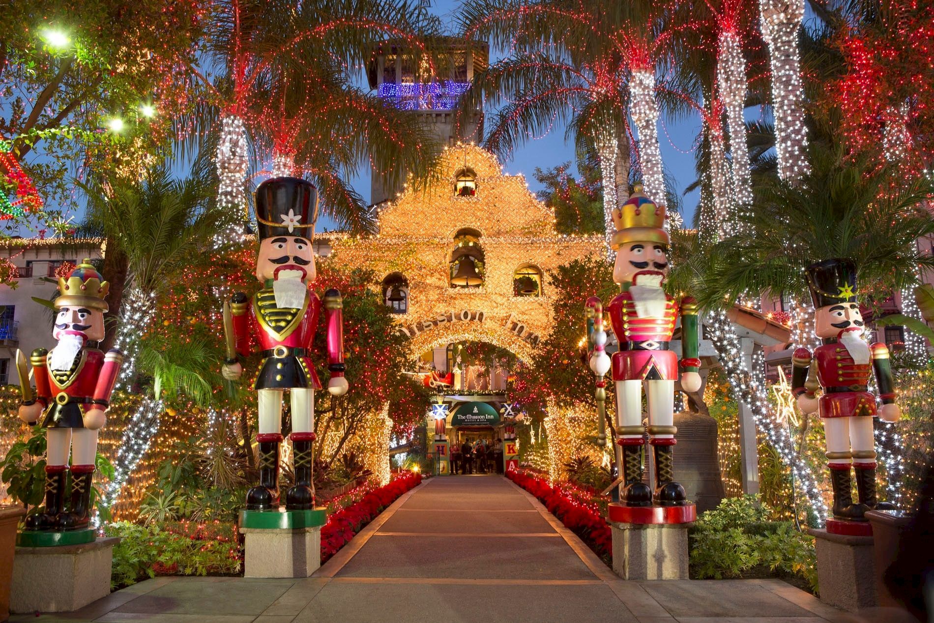 A festive holiday display with towering nutcracker soldiers, palm trees covered in red, gold, and white lights, and a glowing path.