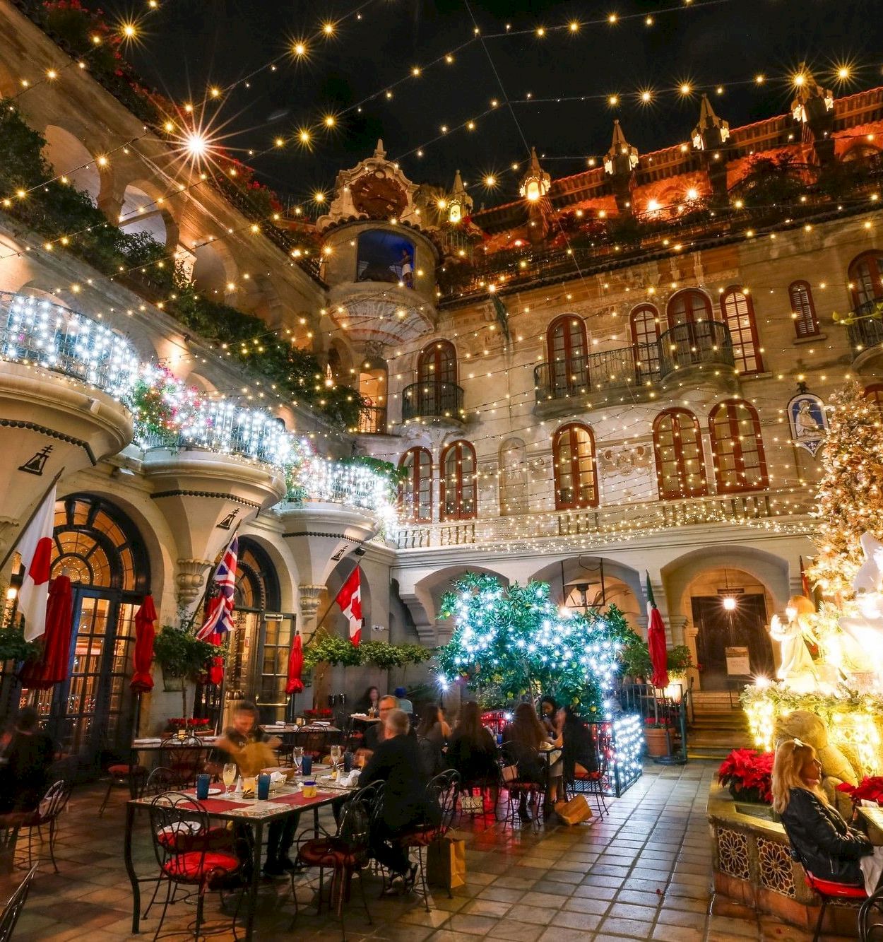 A festive hotel courtyard at night with warm string lights, hanging lanterns, flags, a carousel-like fountain, and diners celebrating.
