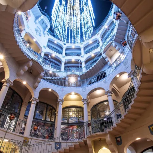 Inside a grand circular atrium with curved balconies and a domed ceiling, glowing with blue-white hanging lights and warm lamps.