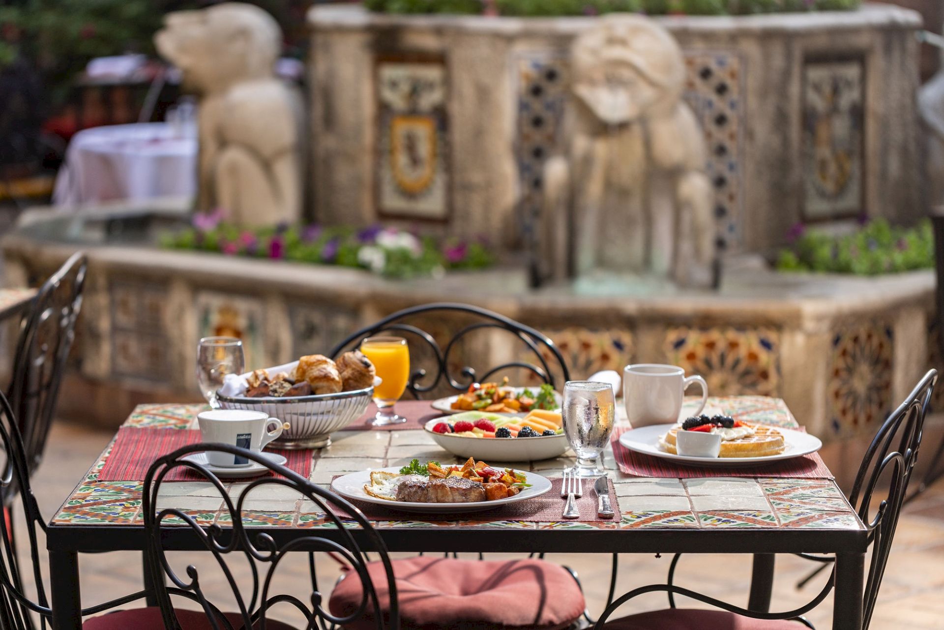 An outdoor breakfast table set with plates, fruit, pastries, juice, and coffee in front of a carved stone garden fountain.