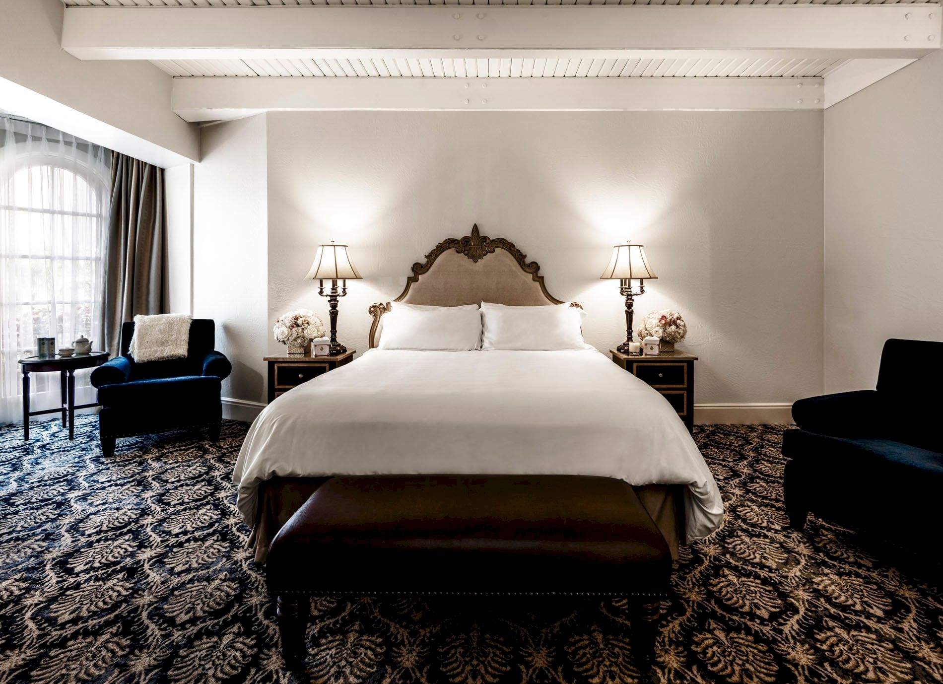 A luxurious hotel bedroom with a carved headboard bed, two lamps, nightstands, curtains, and a patterned carpet.