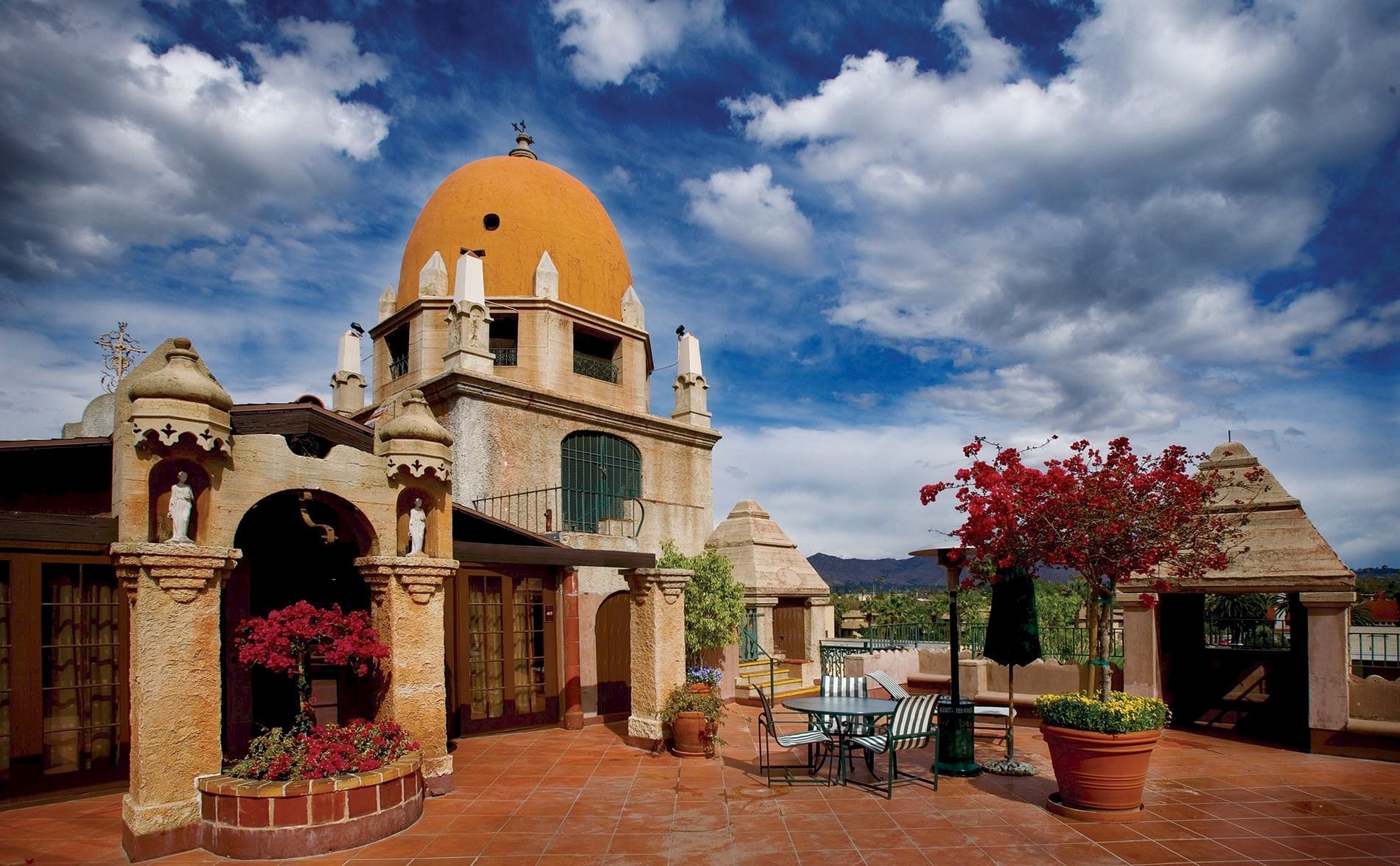 A yellow-domed church in a sunny courtyard, with red flowers and patio seating beneath dramatic blue clouds.
