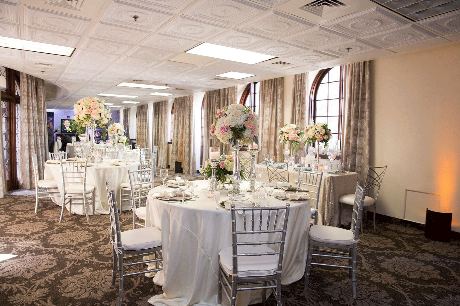 An elegant event hall set for a wedding or banquet, with round tables, silver chairs, and tall floral centerpieces by tall windows.