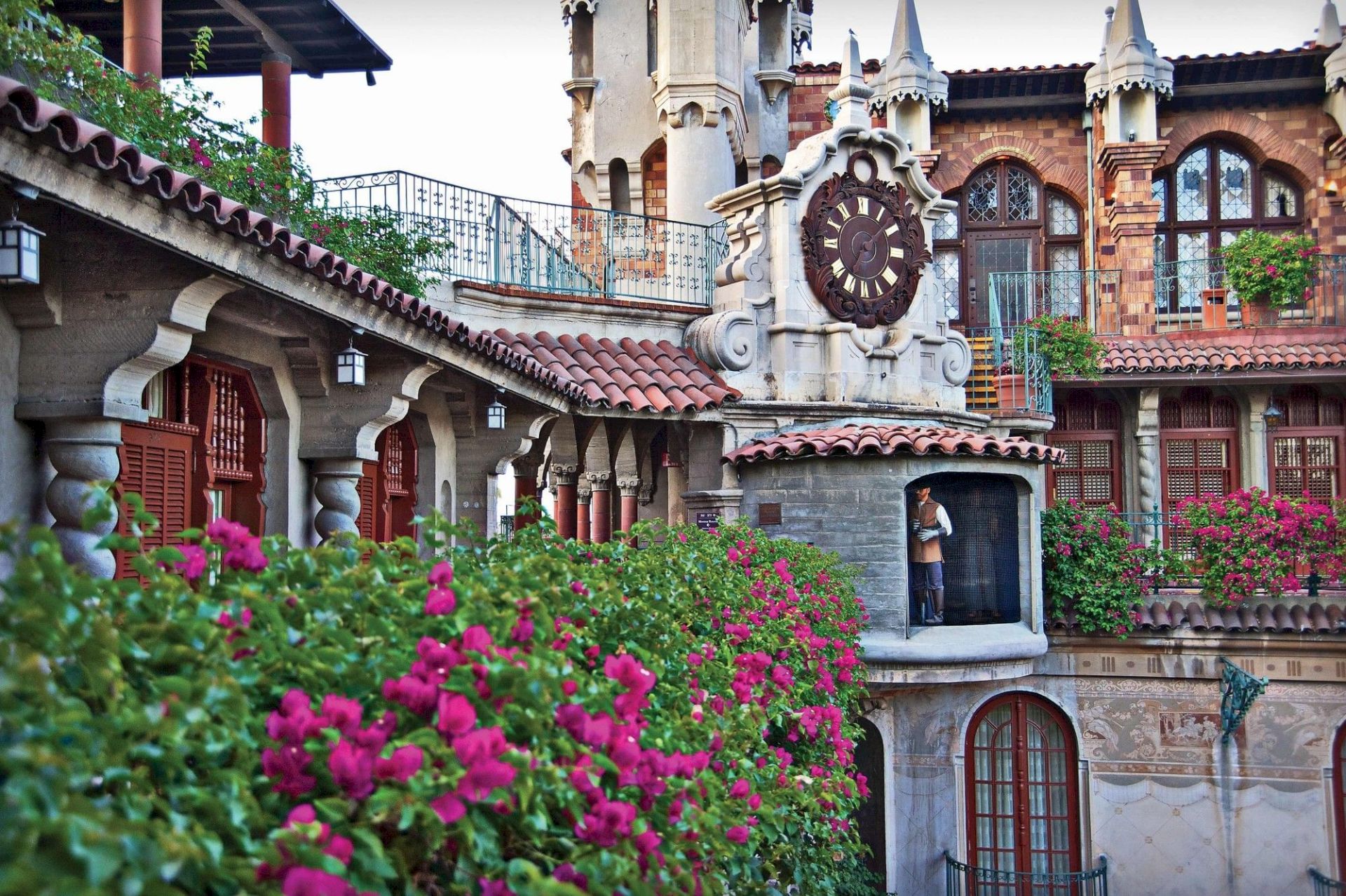 An ornate stucco clock tower with Roman numerals stands beside arched windows, framed by lush pink flowers in the foreground.