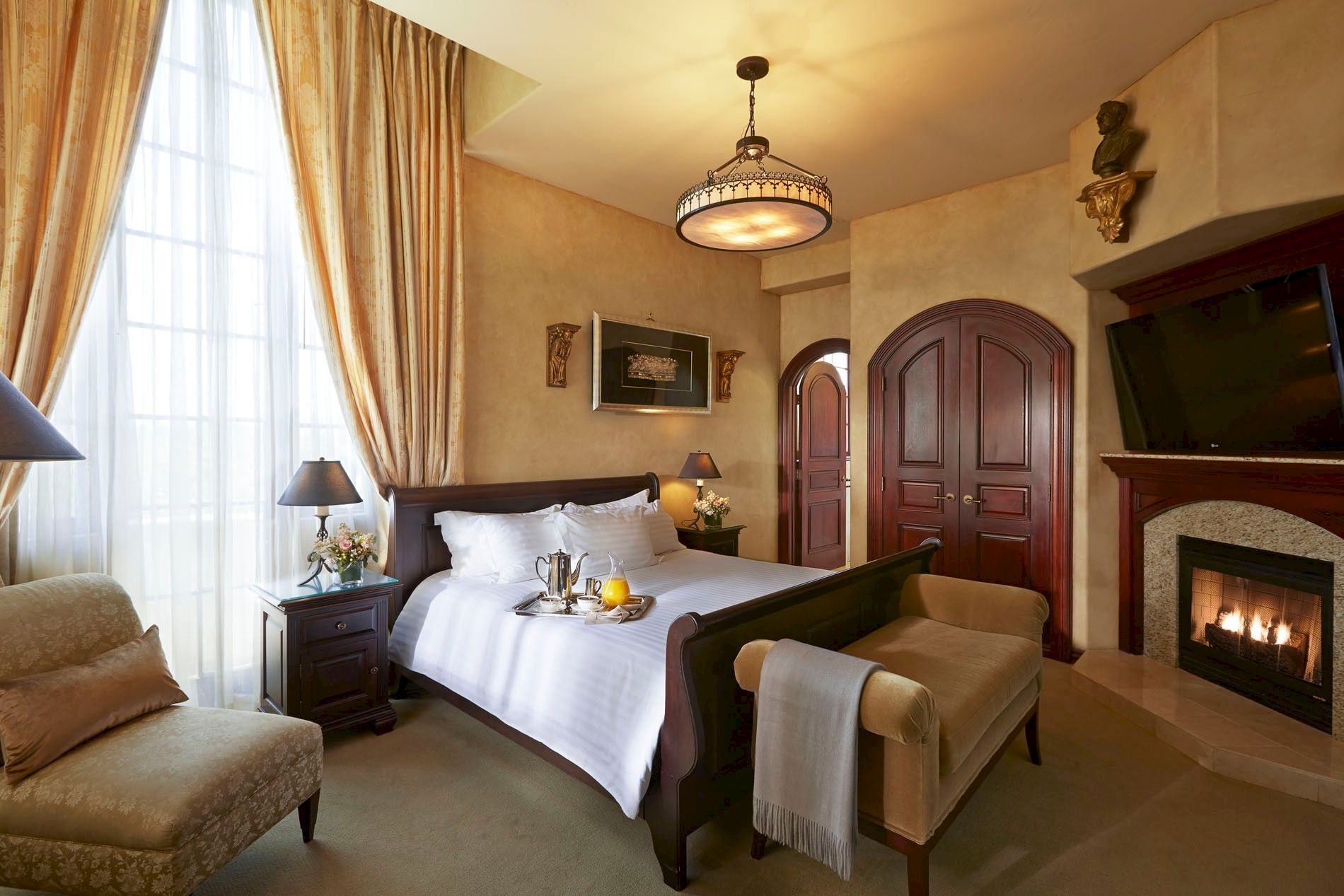 A luxurious hotel-style bedroom with a canopy bed, ornate chandelier, fireplace, and curtained windows, creating a warm, elegant atmosphere.