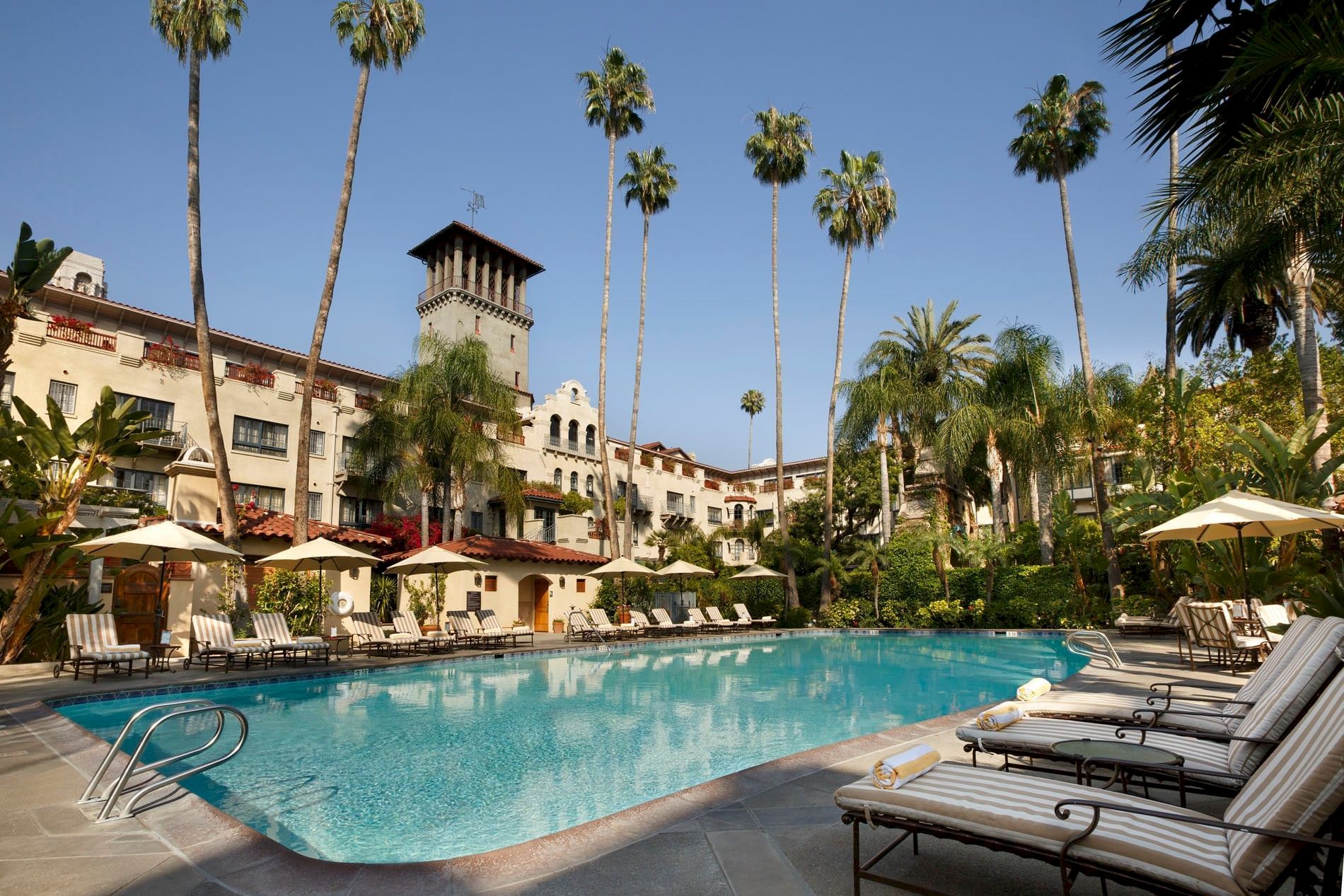 A sunny hotel courtyard with a blue swimming pool, striped loungers, umbrellas, and tall palm trees.