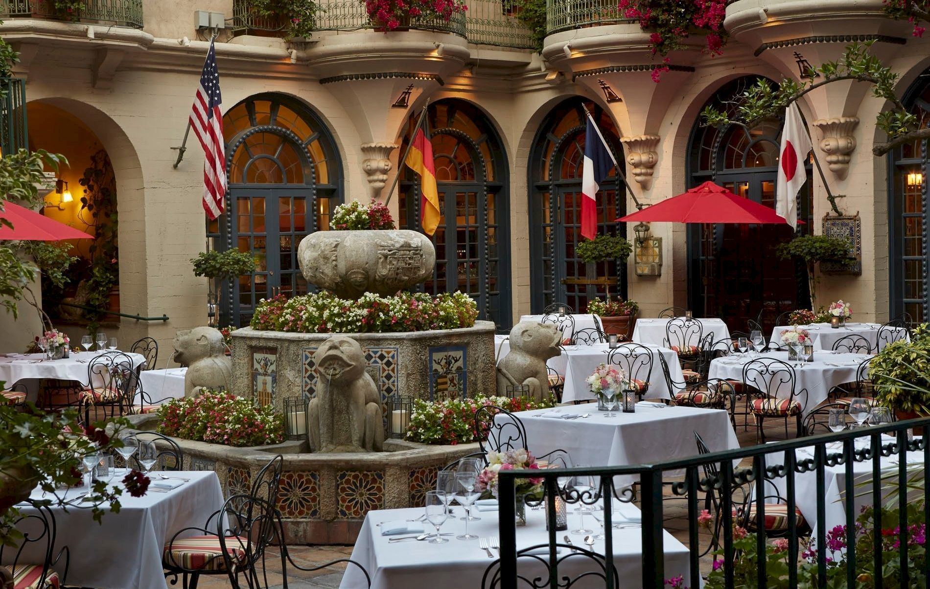 An elegant outdoor courtyard dining setup with white tablecloths, blooming flowers, and several flag-topped arches.