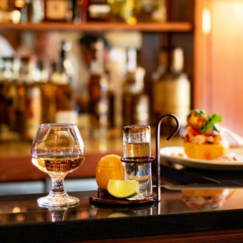A warmly lit bar counter with a glass of amber drink, a cocktail shaker and lime, plus a plated appetizer in the background.