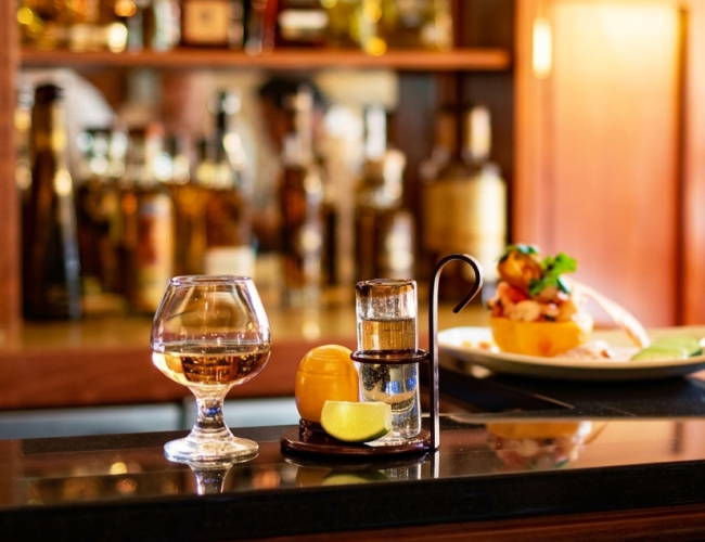 A warmly lit bar counter with a glass of amber drink, a cocktail shaker and lime, plus a plated appetizer in the background.
