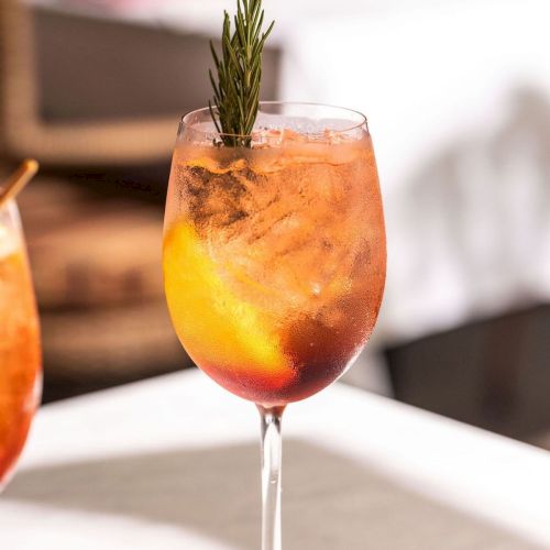 A rosemary-garnished orange cocktail in a stemmed glass, with icy condensation and vibrant yellow-red tones.