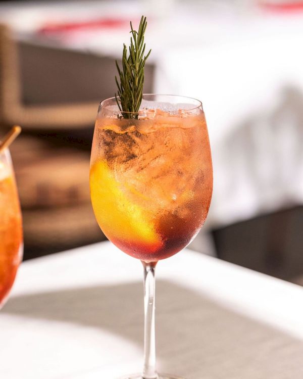 A rosemary-garnished orange cocktail in a stemmed glass, with icy condensation and vibrant yellow-red tones.