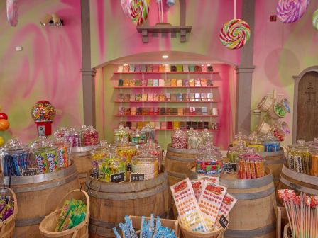 A colorful candy shop with jars of sweets on wooden barrels, lollipops hanging overhead, and shelves of packaged candy.
