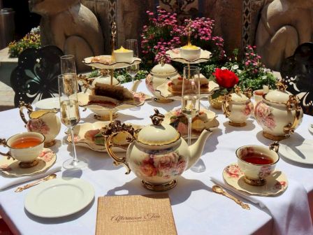 An ornate outdoor afternoon tea setup with floral displays, red umbrellas, a stone fountain, and a decorated teapot and cups.