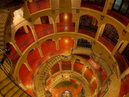 A grand multi-level atrium inside a historic building, with red tiled floors, arched balconies, and a central statue.