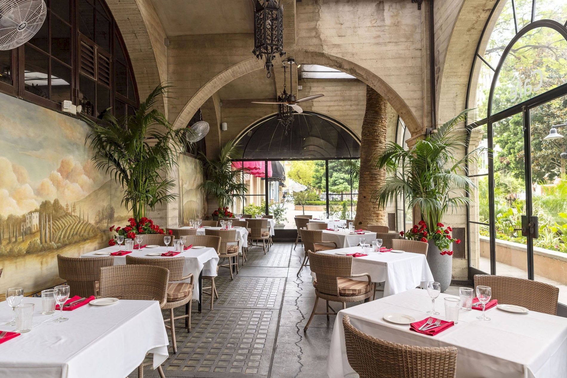 A stylish indoor dining terrace with set tables, palms, arched stone walls, chandelier lighting, and large windows by a mural scene.