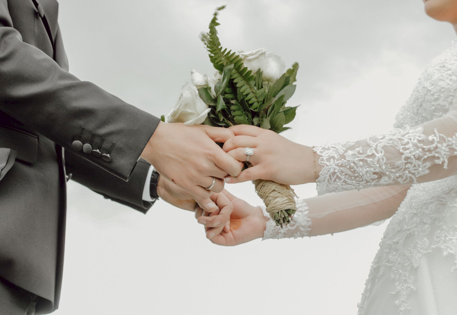A groom in a suit and a bride in lace hold a bouquet while showing wedding rings in a tender, wedding-day moment.