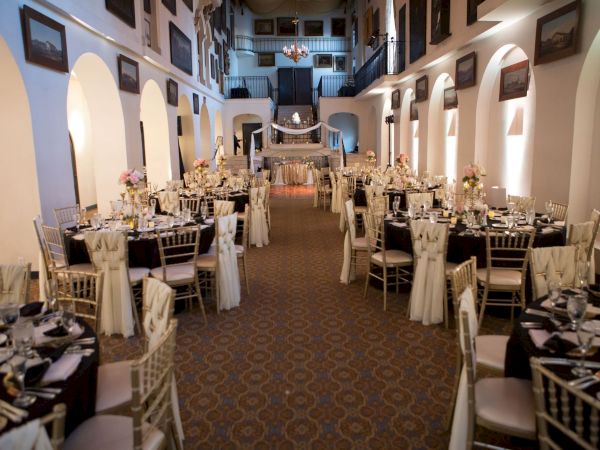 Elegant wedding reception hall set with banquet tables, candles, flowers, and chandeliers in a vaulted interior.