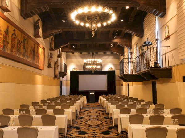 An elegant banquet hall with chandeliers, set tables, and a stage with a screen, framed by ornate balconies and statues.
