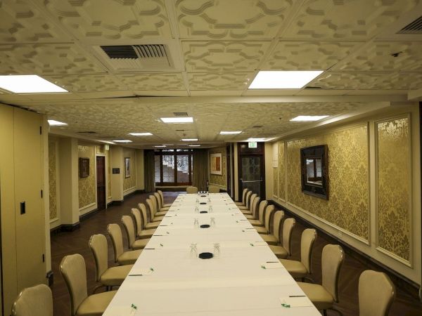 A formal conference room with a long white table, beige chairs, patterned wallpaper, and ornate ceiling lights and trim.
