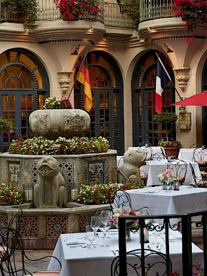 An elegant outdoor courtyard dining set with white tables, red umbrellas, floral vines, and US/Germany/Japan flags on arches, welcoming guests.