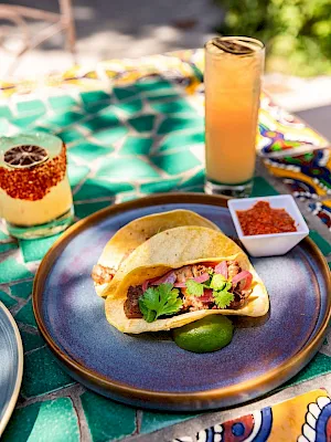 A plate of tacos with cilantro and onions on a colorful table, plus iced drinks and salsa bowls for a sunny meal.