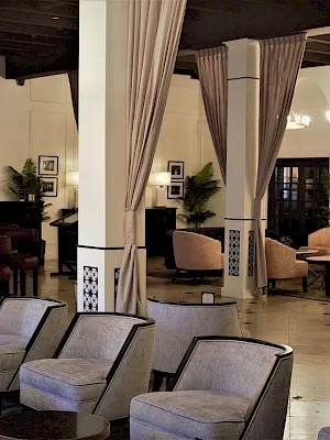 A luxury hotel lounge with upholstered armchairs, tiled floors, elegant draped curtains, and warm chandeliers under dark beams.