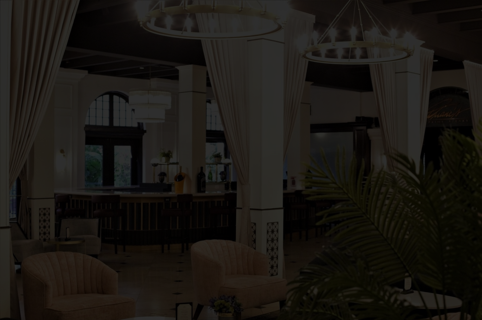 Luxurious hotel lounge with tall curtains, multiple hanging chandeliers, a bar counter, sofas, and plants.