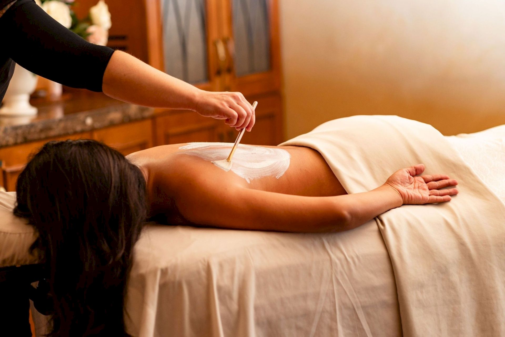 A masseuse applies cream with a spatula during a relaxing back massage on a client lying on a bed.