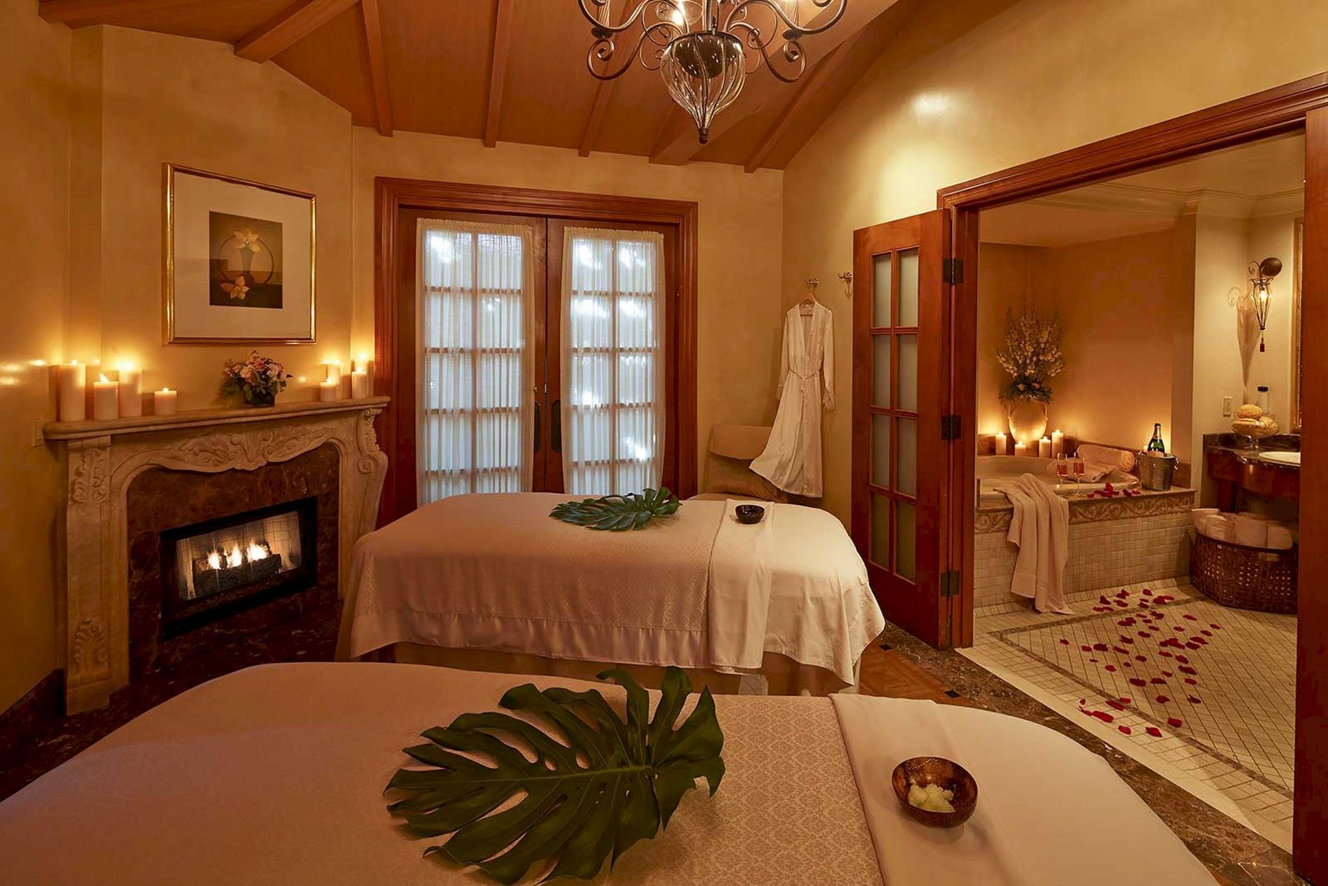 A romantic spa suite with a four-poster bed, lit candles, flower petals, fireplace, and a chandelier over ornate decor.
