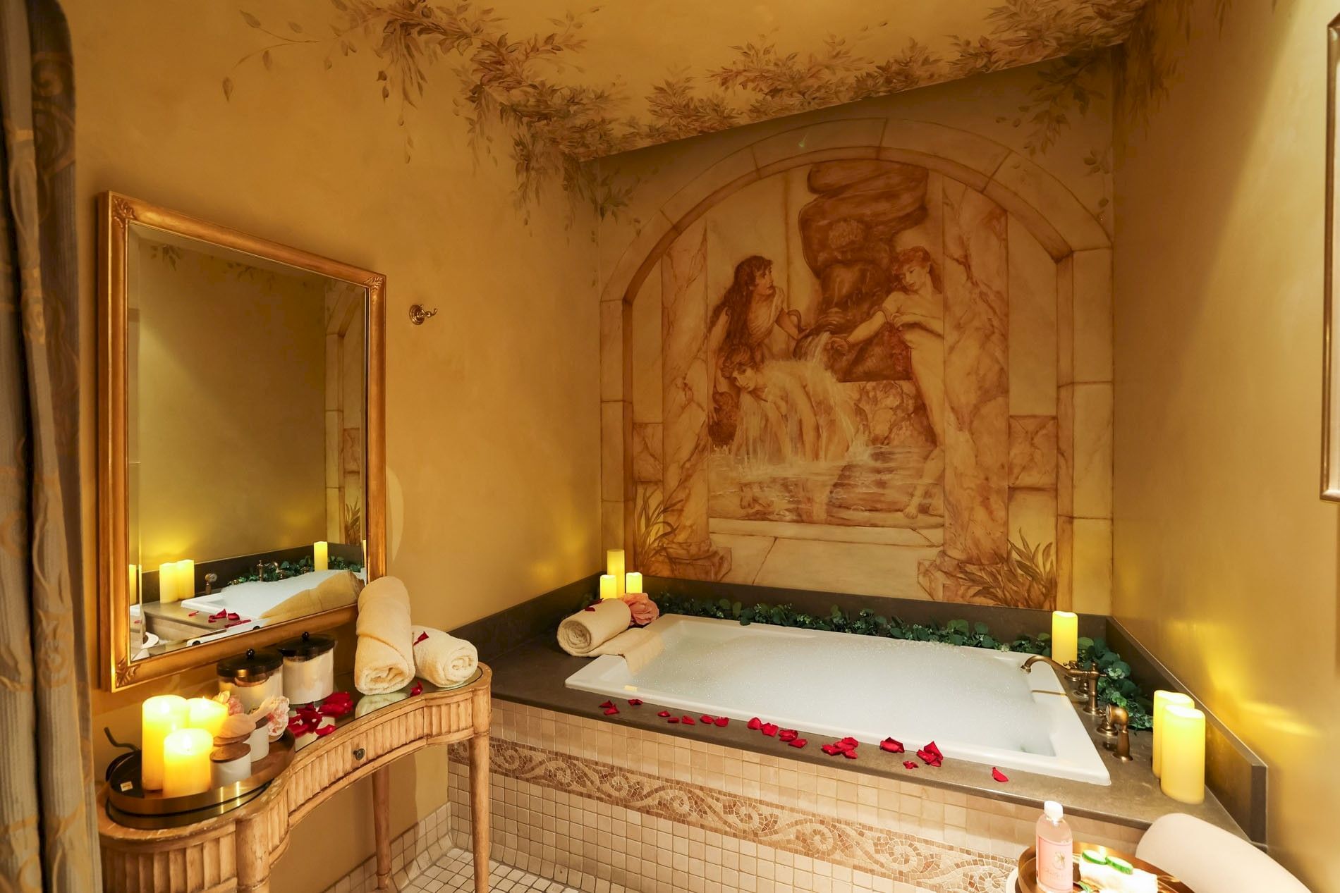 A candlelit spa bathroom with a large bathtub, rose petals, folded towels, and a decorative mural on the wall.