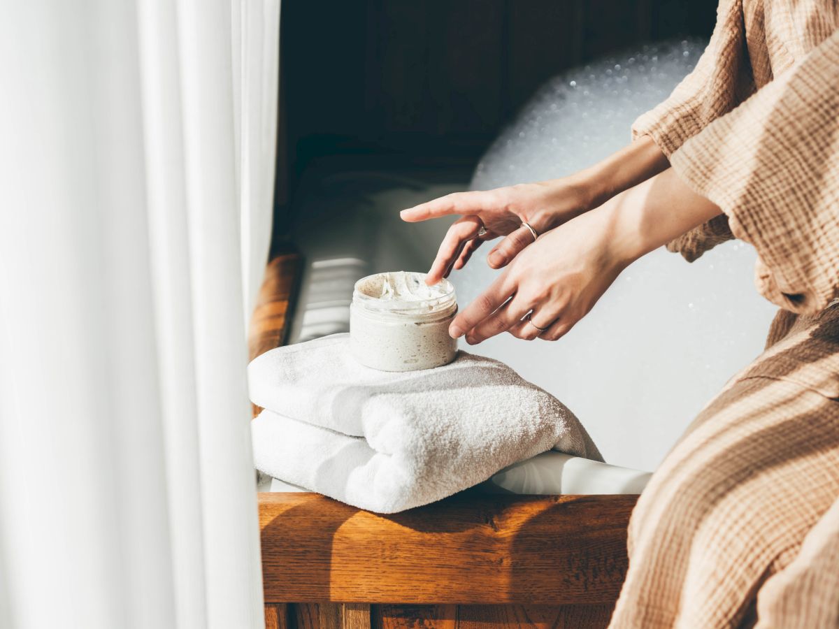 A person scoops lotion from a small jar on a towel, likely prepping for skincare in a bright bathroom.