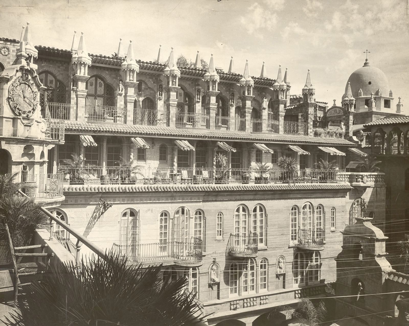 A vintage view shows an ornate stone palace with many archways, rooftop spires, and a large dome under bright sky.