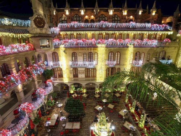 A grand hotel courtyard festooned with red, white, and golden holiday lights and glowing decorative arches at night.