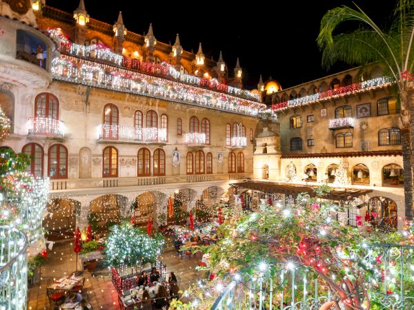 A night courtyard of historic buildings wrapped in bright Christmas lights, with a palm tree and people dining below.