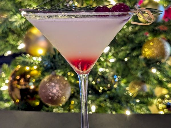 A festive cocktail in a martini glass, topped with dark cherry garnish, with Christmas lights and ornaments blurred in the background.