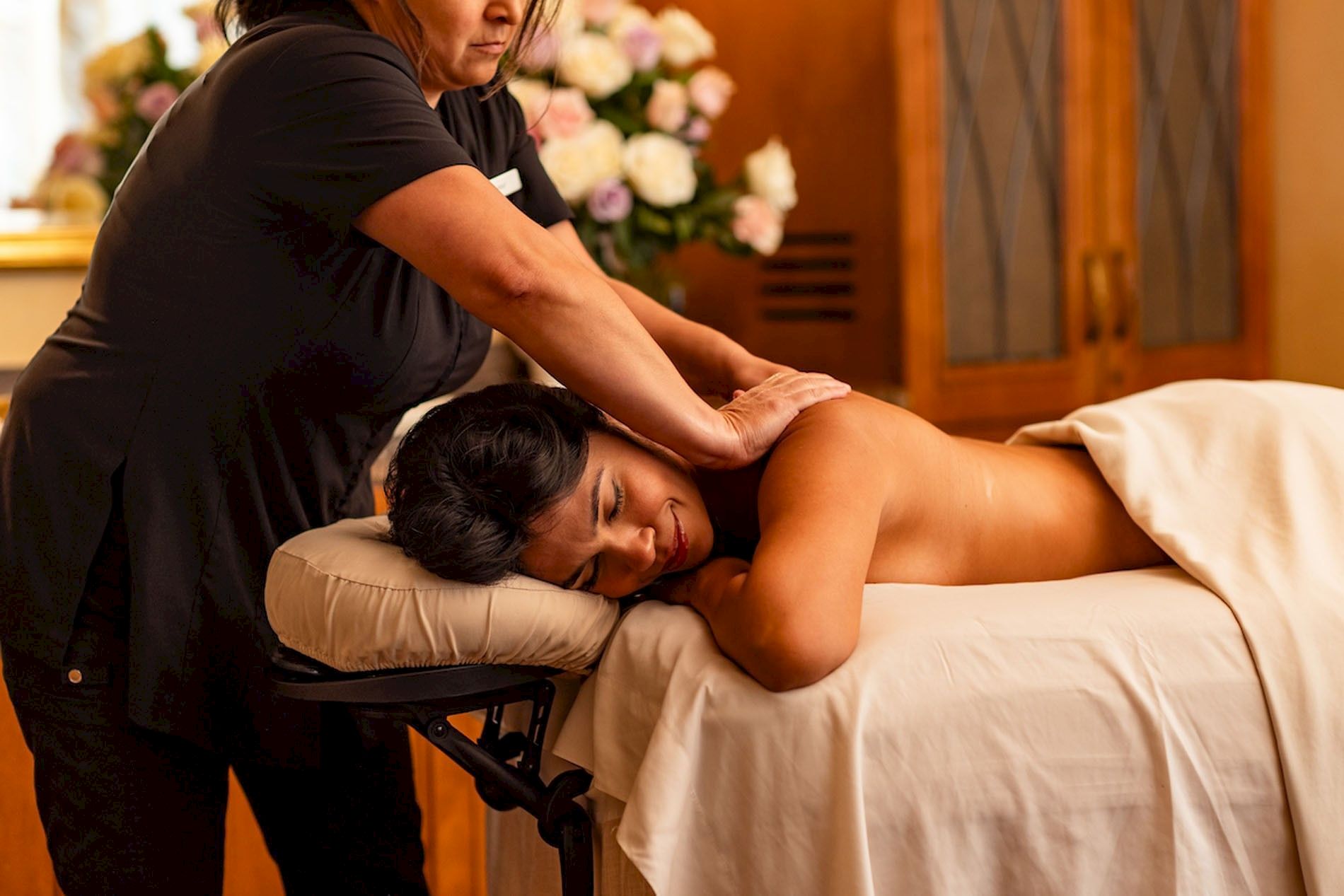 A therapist performs a body massage on a client lying on a treatment table in a warm, flower-filled room.