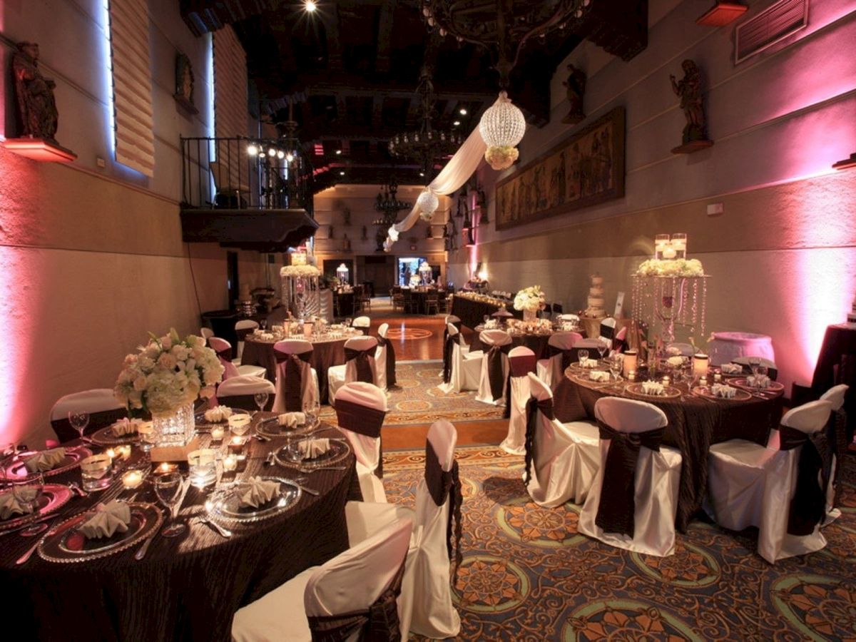 A lavish banquet hall set for a celebration, with round tables, white chair covers, floral centerpieces, chandeliers, and pink stage lighting.