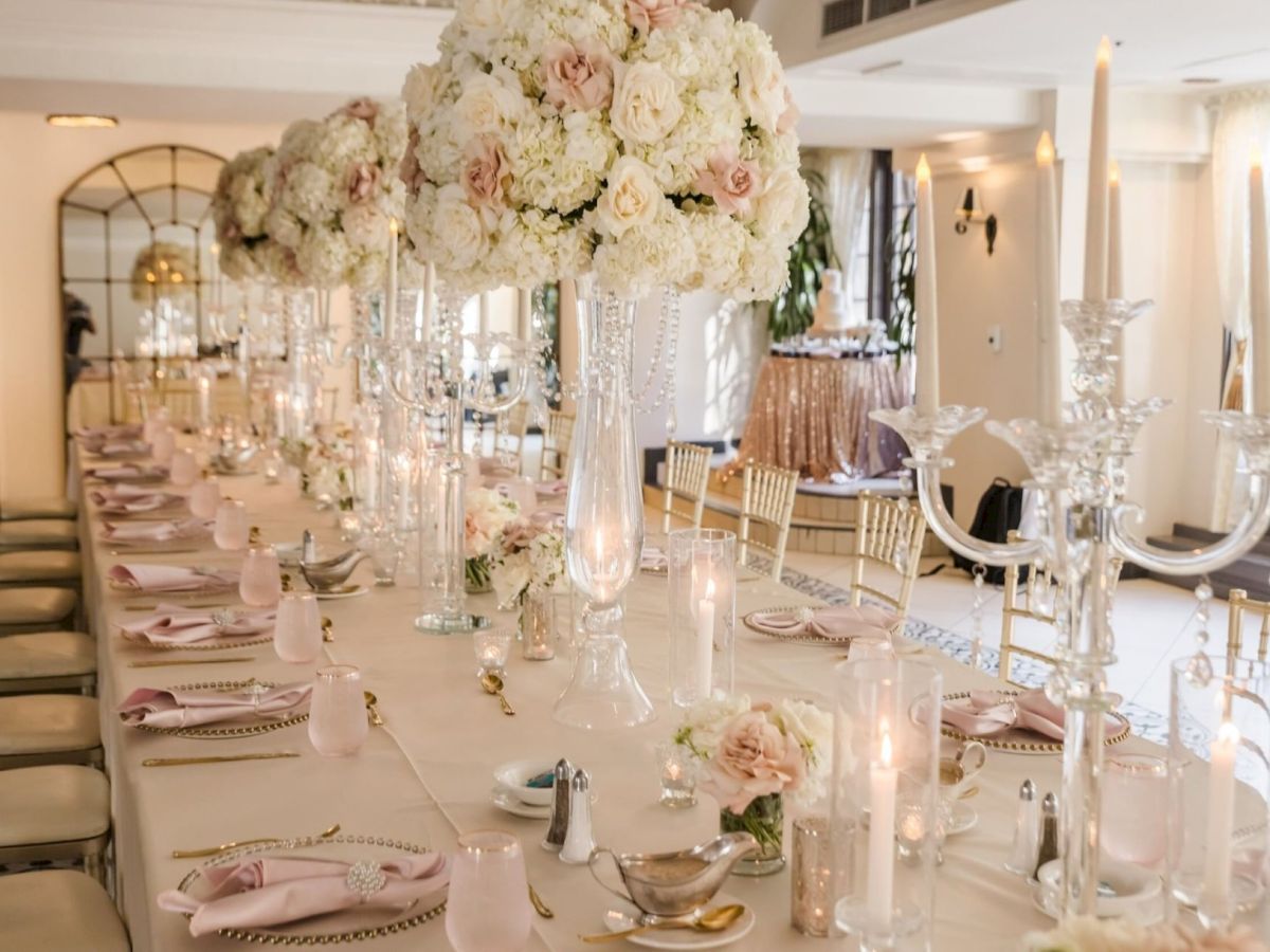 A lavish banquet table set for a wedding, with tall white floral centerpieces, crystal glassware, gold accents, and lit candles.
