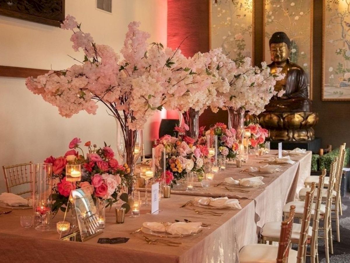 An elegant banquet table is decorated with pink cherry-blossom trees, candles, flowers, glassware, and ornate chairs, with a golden Buddha statue in the background.