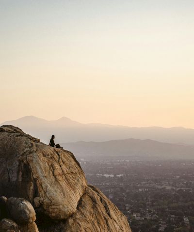 A hiker stands on a rocky hilltop, gazing over a distant city and misty mountains at sunset.