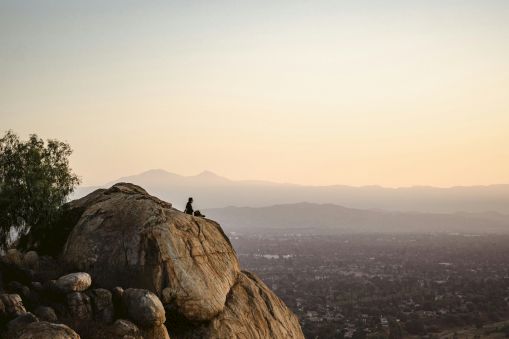 A hiker stands on a rocky hilltop, gazing over a distant city and misty mountains at sunset.