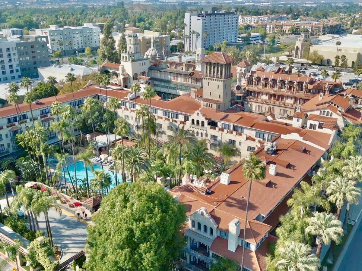 Aerial view of a resort complex with red-tiled roofs, palm trees, courtyards, and a swimming pool under bright sun.