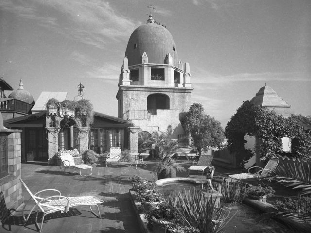 A vintage courtyard scene with lounge chairs, lush plants, and a domed church-like building under a cloudy sky.