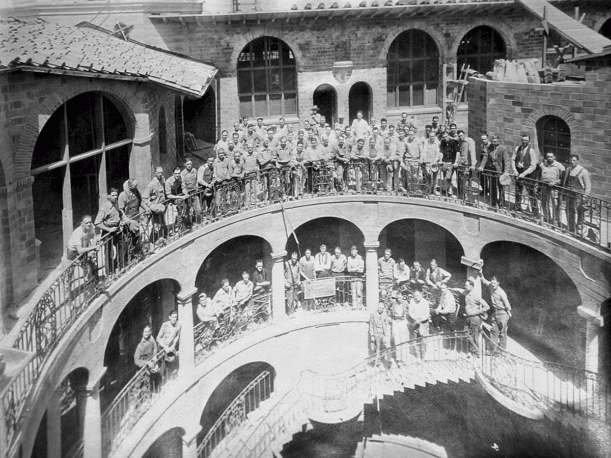 Vintage photo of a large band lined along a curved courtyard bridge with guards/crew in an old stone facility, watching.